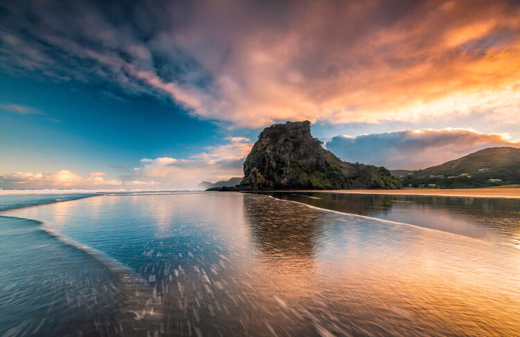 Piha Beach at sunset with Lion Rock and golden reflections on wet sand